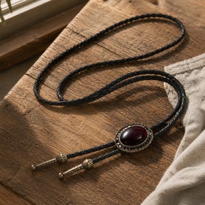 May include: A black bolo tie with a braided leather cord. The tie features a dark red oval stone set in a decorative bronze-colored metal frame. The tips of the tie are metallic.