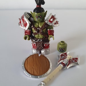 Könnte beinhalten: Eine grüne Ork-Minifigur mit schwarzem Pferdeschwanz, die eine rot-silberne Rüstung mit Schulterpanzern trägt. Neben der Figur liegen ein zusätzlicher grüner Kopf, ein runder brauner Schild mit silbernem Rand und eine Doppelaxt mit weißem Griff.
