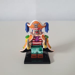Könnte beinhalten: Spielzeugfigur eines Piratencharakters mit clownartigem Gesicht, der einen Dreispitzhut mit Totenkopf-Emblem trägt. Die Figur hat eine rote Nase, ein gestreiftes Hemd und hält zwei Schwerter. Die Figur steht auf einem schwarzen Sockel.