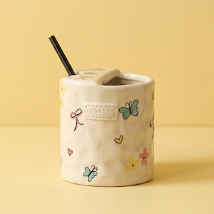 Pode incluir: Uma caneca de cerâmica creme com um canudo preto. A caneca é decorada com borboletas, flores e corações coloridos. O texto "coffee time" está impresso na lateral. A caneca tem um formato único com um topo curvo.