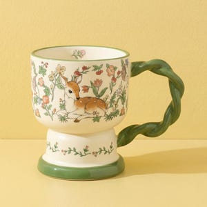 May include: A ceramic mug with a white base and a green handle and base. The mug features a floral and deer design. The handle is a twisted green vine shape. The mug is on a yellow background.