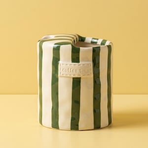 Op de afbeelding: Een keramische container met verticale groene strepen op een crèmekleurige achtergrond. De container heeft een opgerolde bovenkant en een klein rechthoekig label met de tekst "coffee time". De container staat op een gele ondergrond.