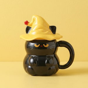 Puede incluir: Una taza de cerámica blanca con asa negra y el texto "But First, Coffee" en negrita negra. La taza se encuentra sobre un fondo borroso, resaltando su diseño limpio.