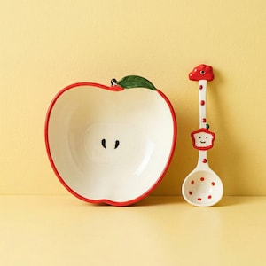 Può includere: Una ciotola in ceramica a forma di mela tagliata, con bordo rosso e foglia verde. Un cucchiaio abbinato con un design a mela rossa e pois rossi è accanto alla ciotola. Il tutto è su sfondo giallo.