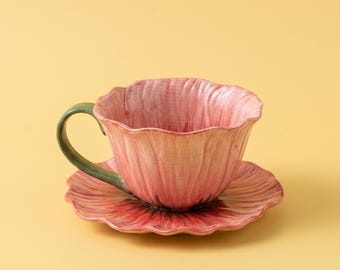 Könnte beinhalten: Ein Keramik-Tassenset mit Untertasse in Form einer blühenden Blume. Tasse und Untertasse zeigen ein rosa, blütenblattartiges Design mit gewelltem Rand und einem grünen Henkel, der an einen Stiel erinnert. Die Oberfläche hat ein glänzendes Finish.