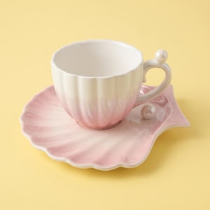 May include: A ceramic teacup and saucer set featuring a scalloped shell design. The cup has a white interior and a pink gradient exterior. The saucer is shaped like a large seashell in a soft pink hue, accented with a small white pearl-like bead.