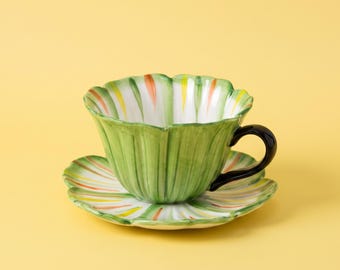 May include: A ceramic teacup and saucer set featuring a scalloped, flower-like shape. The exterior and interior are painted with vertical green stripes and accents of yellow and orange. The teacup includes a solid black, curved handle.