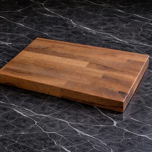 Puede incluir: Una tabla de cortar rectangular de madera con un patrón de vetas de madera natural. La tabla está hecha de múltiples piezas de madera unidas. La tabla de cortar se muestra sobre una superficie de mármol negro.