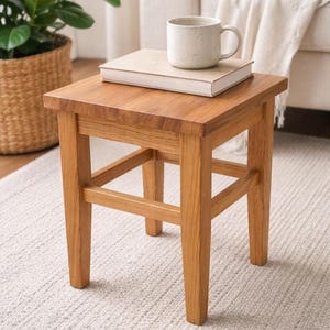 Puede incluir: Un pequeño taburete cuadrado de madera con acabado natural. Un libro y una taza de cerámica moteada están encima. El taburete tiene cuatro patas y una barra transversal. El taburete mide 38 cm de alto.