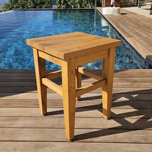 Può includere: Uno sgabello quadrato in legno con una finitura naturale color marrone chiaro, dotato di quattro gambe dritte collegate da barre di supporto orizzontali. Lo sgabello presenta una superficie superiore piatta con venature del legno visibili.