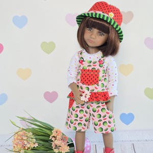 Könnte beinhalten: Eine Puppe in einem Outfit zum Thema Wassermelone, einschließlich Hut, Latzhose und Shorts. Der Hut ist rot mit schwarzen Samen und einem grünen Rand. Die Latzhose und die Shorts haben einen rosa Hintergrund mit Wassermelonenscheiben. Die Puppe trägt rote Schuhe.