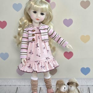 Puede incluir: Una muñeca rubia con dos pequeños pompones rosas en el pelo, vestida con un vestido rosa de flores con dobladillo con volantes y una chaqueta de rayas con tres botones. La muñeca lleva calcetines marrones de lunares y zapatos rosas junto a dos ovejas.
