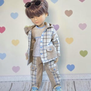May include: A doll with brown hair wearing round sunglasses, a light blue and beige plaid hooded jacket, a light blue and gray t-shirt, matching plaid cargo pants with blue buttons, and light blue and beige sneakers. The text Dollsgarmentlily is visible.