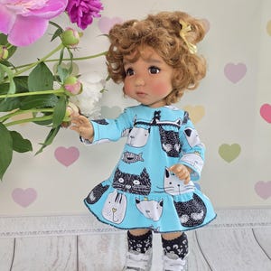 May include: A doll with curly brown hair and a yellow hair clip, wearing a light blue long-sleeve dress with a black and white cat print, black and white patterned socks, and white sneakers. The text Dollsgarmentlily is visible at the top.