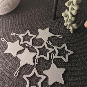 Könnte beinhalten: Eine Sammlung silberner sternförmiger Ornamente mit angebrachten Haken. Einige Sterne sind massiv, andere haben eine offene Kontur. Die Ornamente sind auf einer dunklen, strukturierten Oberfläche angeordnet, was auf ein dekoratives oder handwerkliches Thema hindeutet.
