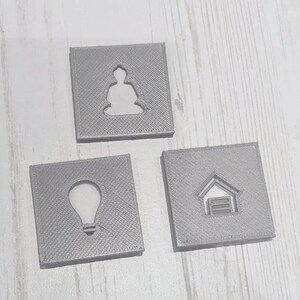 Könnte beinhalten: Drei graue quadratische Kacheln mit ausgeschnittenen Formen: eine sitzende Figur, eine Glühbirne und ein Haus mit Garagentor. Die Kacheln bestehen aus einem strukturierten Material und liegen auf einer hellen Holzoberfläche.