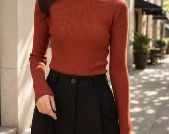 May include: A rust-colored long-sleeve turtleneck sweater with a ribbed knit texture, paired with black trousers featuring a high-waisted design, a single visible button closure at the center, and subtle pleats below the waistband.