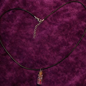 May include: A black cord necklace with a small glass vial pendant. The vial is filled with pink and red decorative elements and sealed with a cork. The background is a deep purple velvet.