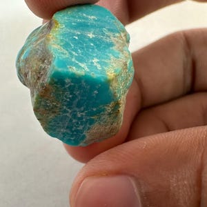 May include: A raw, unpolished turquoise stone featuring a vibrant cyan-blue color with natural brown matrix inclusions. The mineral specimen has an irregular, jagged shape with a textured surface, showcasing organic variations in its blue and brown hues.
