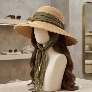 May include: A wide-brimmed straw hat in a natural tan color, featuring a dark olive green fabric band wrapped around the crown. The band extends into two long, tied fabric ribbons that drape down the front, adding a distinct layered texture.