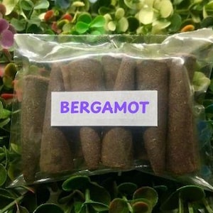 Puede incluir: Una bolsa de plástico transparente llena de varillas de incienso marrones en forma de cono. Una etiqueta blanca con la palabra "BERGAMOT" en letras moradas está adherida al frente. El fondo es una mezcla de plantas artificiales verdes y coloridas.