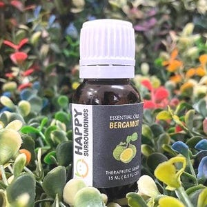 Könnte beinhalten: Eine dunkelbraune Glasflasche mit weißem Deckel, beschriftet mit "Happy Surroundings" und "Essential Oil Bergamot". Das Etikett zeigt eine Abbildung der Bergamotte-Frucht. Die Flasche enthält 15 ml Öl in therapeutischer Qualität, umgeben von farbenfrohem Laub.