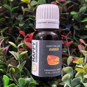 Op de afbeelding: Een kleine, donkere glazen fles amber essentiële olie met een witte dop. Het etiket luidt "Essential Oils AMBER" en "Happy Surroundings". De fles is omgeven door groene en rode bladeren.