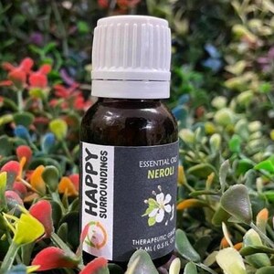 May include: A brown glass bottle with a white cap, labeled "Happy Surroundings Essential Oil Neroli." The label features a floral illustration and the text "Therapeutic Grade 15 ml (0.5 FL OZ)." The bottle is surrounded by colorful plants.