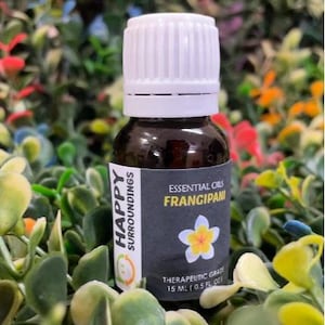 Può includere: Una bottiglia di vetro marrone con tappo bianco, etichettata "Happy Surroundings" e "Essential Oils Frangipani". L'etichetta presenta una grafica floreale bianca e gialla. La bottiglia contiene 15 ml di olio di grado terapeutico.