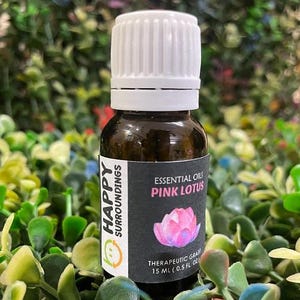 以下が含まれることがあります： 白いキャップが付いた茶色のガラス瓶で、「Pink Lotus」エッセンシャルオイルと表示されています。ラベルにはピンクの蓮の花のイラストと「Happy Surroundings」の文字が描かれています。ボトルには15 mlの治療グレードオイルが含まれています。