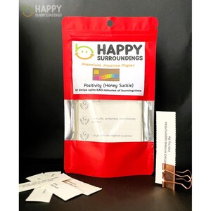以下が含まれることがあります： Happy Surroundingsのプレミアムお香紙の赤いポーチ。パッケージには、白いお香ストリップを示す透明な窓があります。パッケージのテキストには「Positivity（Honey Suckle）」と「32 Strips upto 640 minutes of burning time」と書かれています。
