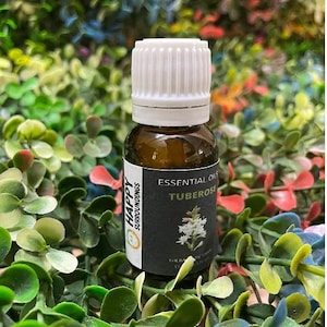 May include: A small amber glass bottle with a white cap. The label reads "Essential Oils Tuberose" with a floral illustration. The bottle is surrounded by green and red foliage, suggesting a natural product.