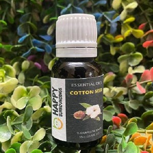 Pode incluir: Uma garrafa de vidro escuro de óleo essencial "Cotton Seed" com tampa branca. O rótulo apresenta uma ilustração de planta de algodão e as palavras "Happy Surroundings". A garrafa é cercada por folhagens verdes e coloridas.