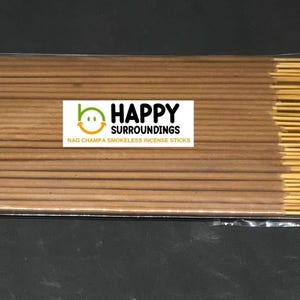 Może przedstawiać: Opakowanie bezdymnych patyczków do kadzidła Nag Champa. Patyczki są w kolorze jasnobrązowym i są zebrane w pakiety. Opakowanie zawiera białą etykietę z napisem "HAPPY SURROUNDINGS" i logo uśmiechniętej twarzy.