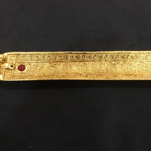 May include: A gold-colored incense holder with intricate embossed patterns and a small red gem. The rectangular tray features a detailed design and a small decorative element at one end. The holder is designed to hold incense sticks.