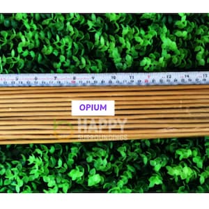 Può includere: Un fascio di sottili bastoncini di legno marrone chiaro, possibilmente incenso, è disposto su uno sfondo di fogliame verde. Un metro a nastro è posto sopra i bastoncini. La parola "OPIUM" è visualizzata su un'etichetta bianca.
