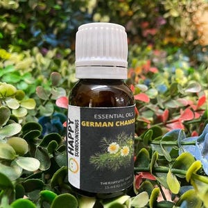 Op de afbeelding: Een bruine glazen fles met een witte dop, met het opschrift "Essential Oils German Chamomile". Het etiket toont een tekening van kamillebloemen en de tekst "Happy Surroundings". De fles bevat 15 ml therapeutische olie.