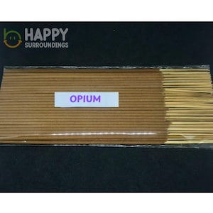 May include: A bundle of brown incense sticks packaged in clear plastic. A white rectangular label is centred on the front with the word OPIUM printed in purple capital letters. The sticks have exposed wooden ends on the right side of the package.