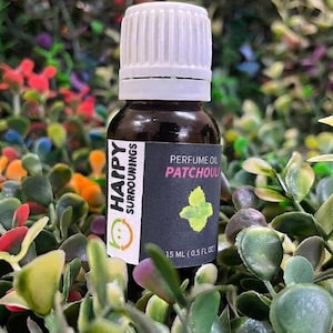 Puede incluir: Un frasco pequeño de vidrio ámbar oscuro con tapa blanca estriada que contiene aceite de perfume. La etiqueta negra presenta el texto Happy Surroundings, Perfume Oil Patchouli y 15 ml (0,5 fl oz) con una pequeña ilustración de hoja verde.