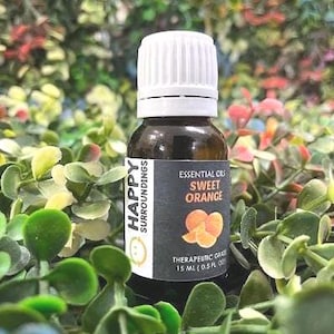 Può includere: Una piccola bottiglia di vetro marrone con tappo bianco, etichettata "Essential Oils Sweet Orange". L'etichetta presenta fette di arancia e il testo "Happy Surroundings". La bottiglia contiene 15 ml di olio di grado terapeutico, circondata da verde.