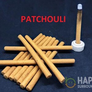 Pode incluir: Uma pilha de varetas de incenso cilíndricas castanho-claras com o texto PATCHOULI em vermelho. Uma vareta acesa está na vertical num pequeno suporte circular branco. O texto HAPPY SURROUNDINGS é visível no canto inferior direito da imagem.