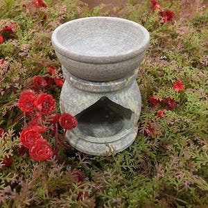 Puede incluir: Un difusor de aceites esenciales de piedra gris con una parte superior en forma de cuenco y una base decorativa. El difusor se encuentra entre vegetación y flores rojas, creando una estética natural. El diseño incluye una pequeña abertura para una vela o fuente de calor.