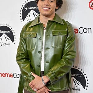 May include: Olive green leather jacket with a button-up front, two chest pockets, and a matching belt. The jacket has white stitching and is paired with brown pants. A silver chain necklace and bracelet complete the look.