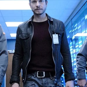 May include: A person wearing a black leather jacket over a burgundy sweater, paired with grey jeans and a black belt. The jacket has a zip closure and a small tag that says "Visitor". The person is in a hallway with blue lighting.