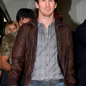 May include: A man wearing a brown leather jacket over a gray plaid shirt and blue jeans. The jacket is zipped up, and the jeans have a distressed look. The man is smiling.