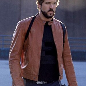 May include: A brown leather jacket with a stand-up collar and a front zip. The jacket is worn over a black top and dark blue trousers. The person is wearing black gloves.