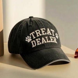 May include: A black baseball cap with a washed-out finish featuring the white embroidered text TREAT DEALER in an arched font, flanked by two small white paw print icons on the front panel above the curved brim of the hat.
