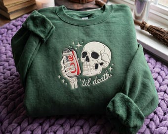 May include: A forest green crewneck sweatshirt featuring an embroidered graphic of a human skull and a skeletal hand holding a red Diet Coke can. The design includes small star accents and the cursive text 'til death.' in white thread.