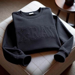 May include: A black long-sleeve crewneck sweatshirt featuring the text X-RAY embroidered in a matching black thread across the chest. The garment has a ribbed collar, ribbed cuffs, and a standard fit, showcasing a monochromatic design throughout.