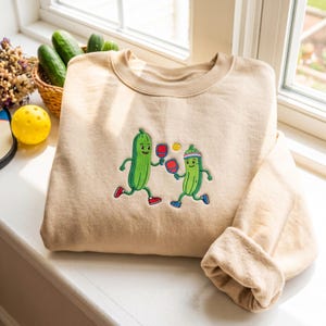 Op de afbeelding: Een beige sweater met ronde hals en een geborduurde afbeelding van twee groene antropomorfe augurken die pickleball spelen. De augurken rennen met rode rackets, waarbij er één een hoofdband en blauwe sneakers draagt en de andere rode sneakers.